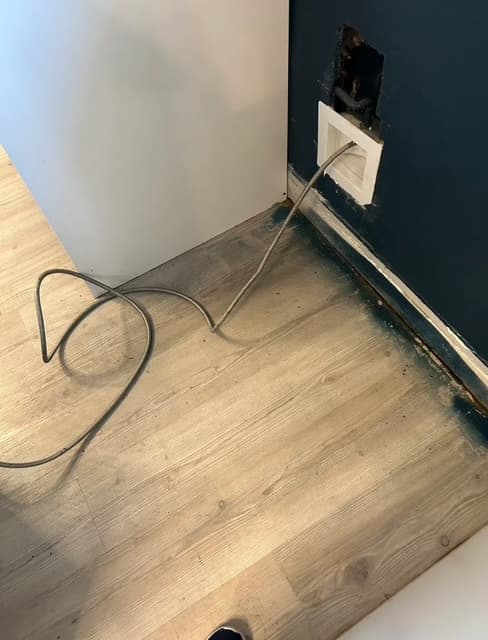 Flexible metal water line extending from a recessed wall box onto light-colored wood-look flooring.