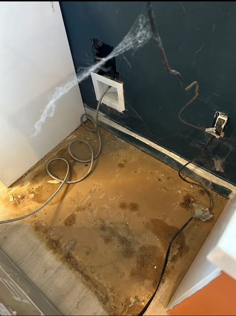 Large brown stain on floor behind an appliance with tangled cables and hanging cobwebs.