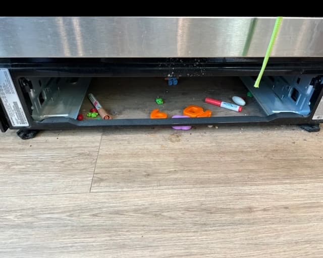 Markers, plastic letters, and small toys scattered on the floor underneath a stainless steel stove.