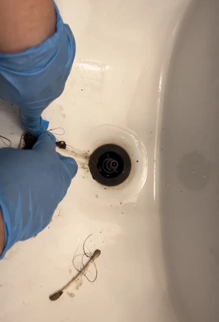Hands in blue gloves removing a hair clog from a white sink drain.