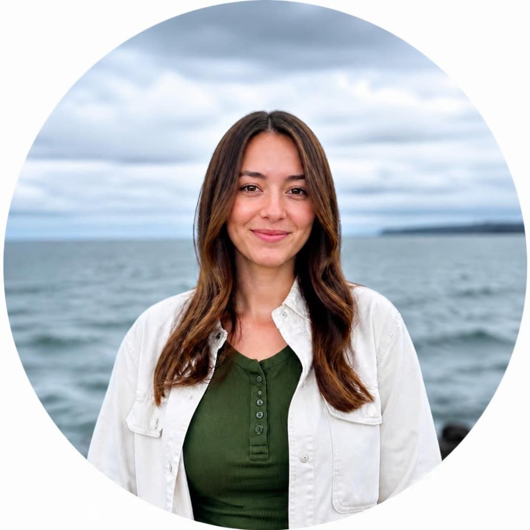 Smiling woman with long brown hair wearing a white jacket, ocean and cloudy sky behind her.