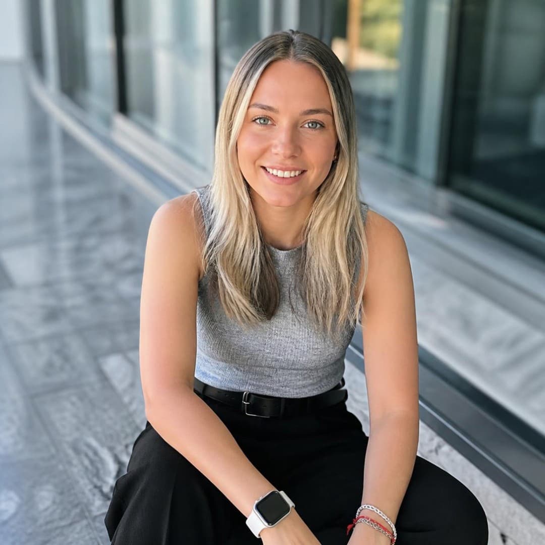 Smiling blonde woman wearing a grey top and black pants sits outside a modern building.
