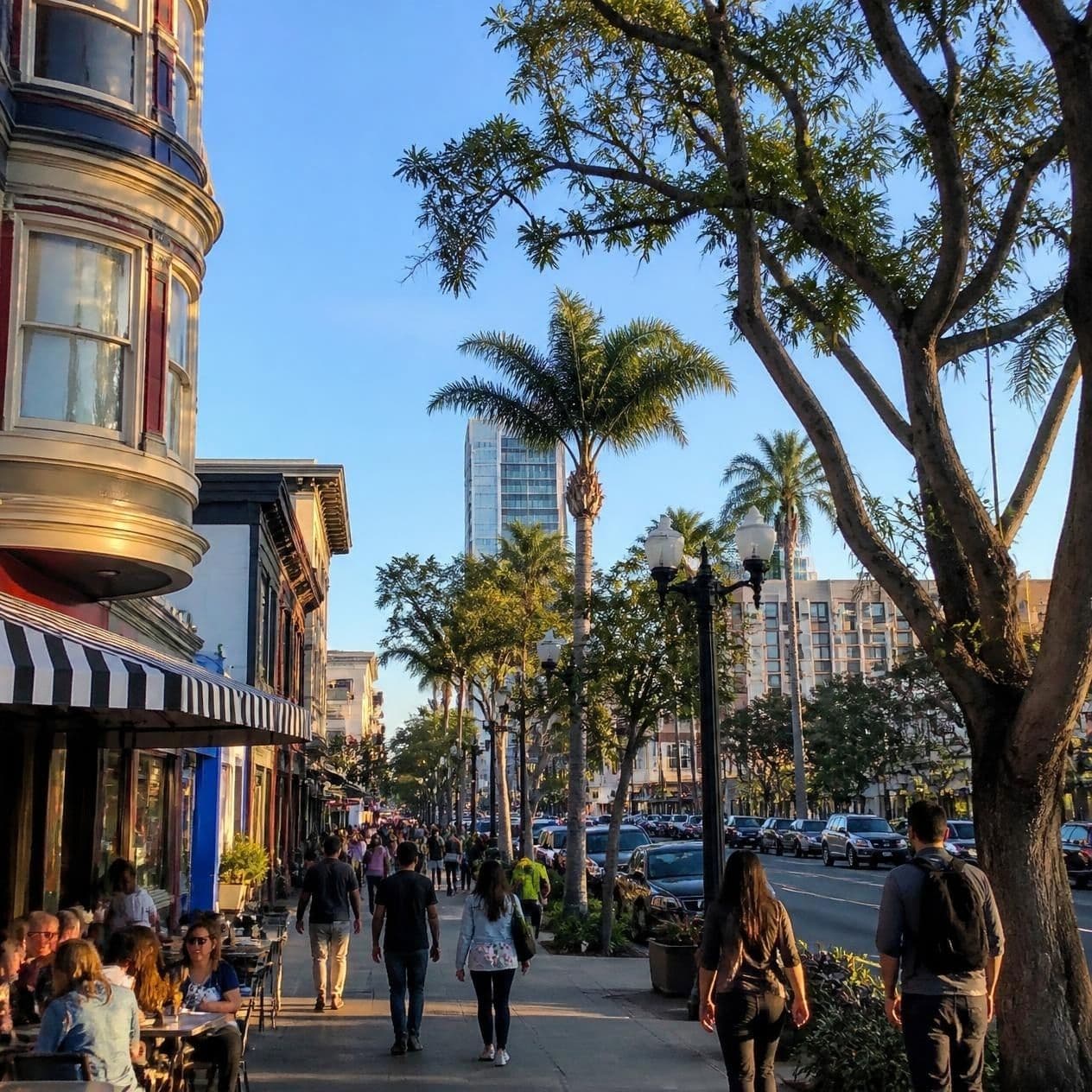 A bustling city sidewalk at sunset with palm trees, pedestrians, and outdoor restaurant seating.