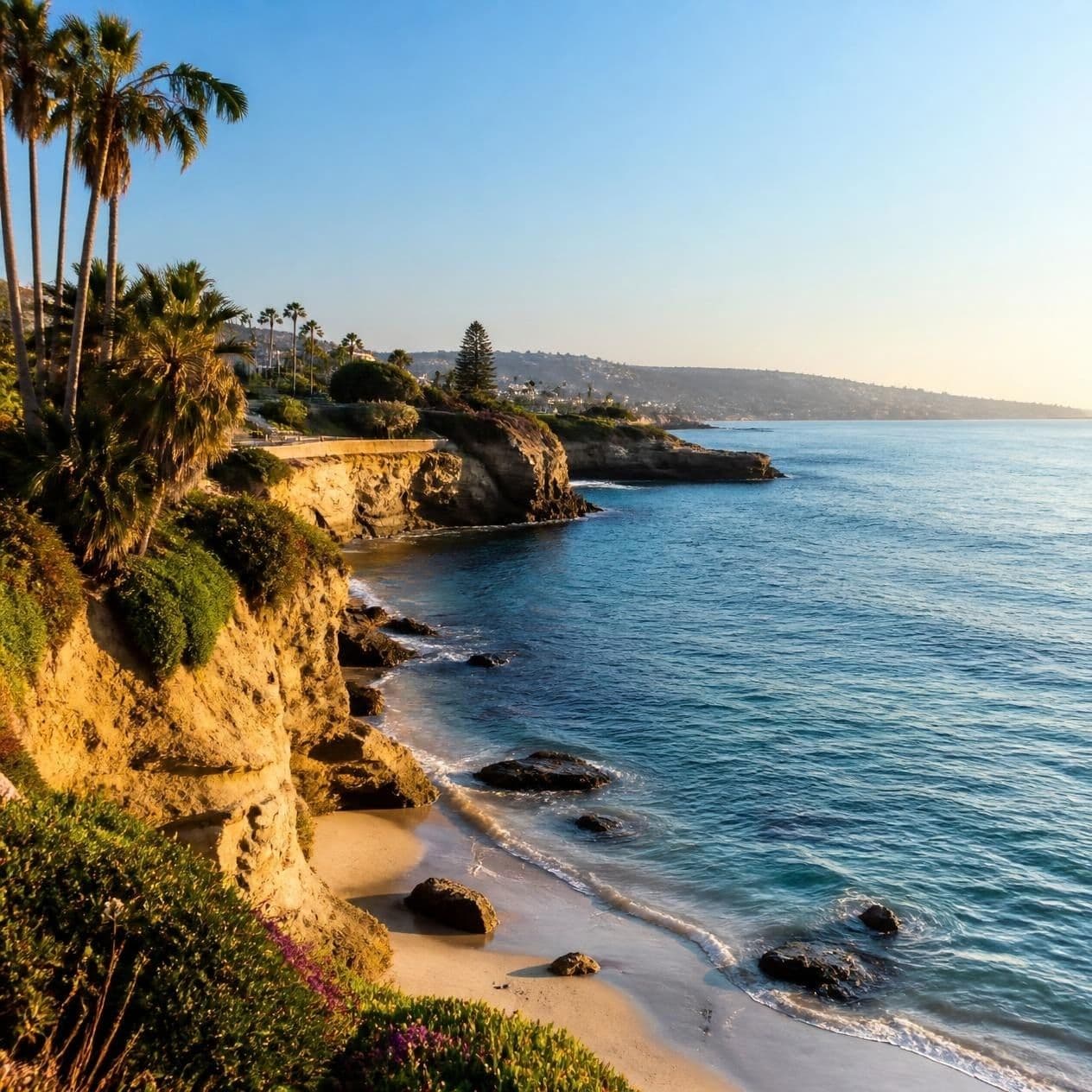Golden sunlight illuminates rocky cliffs and palm trees overlooking a serene blue ocean beach.
