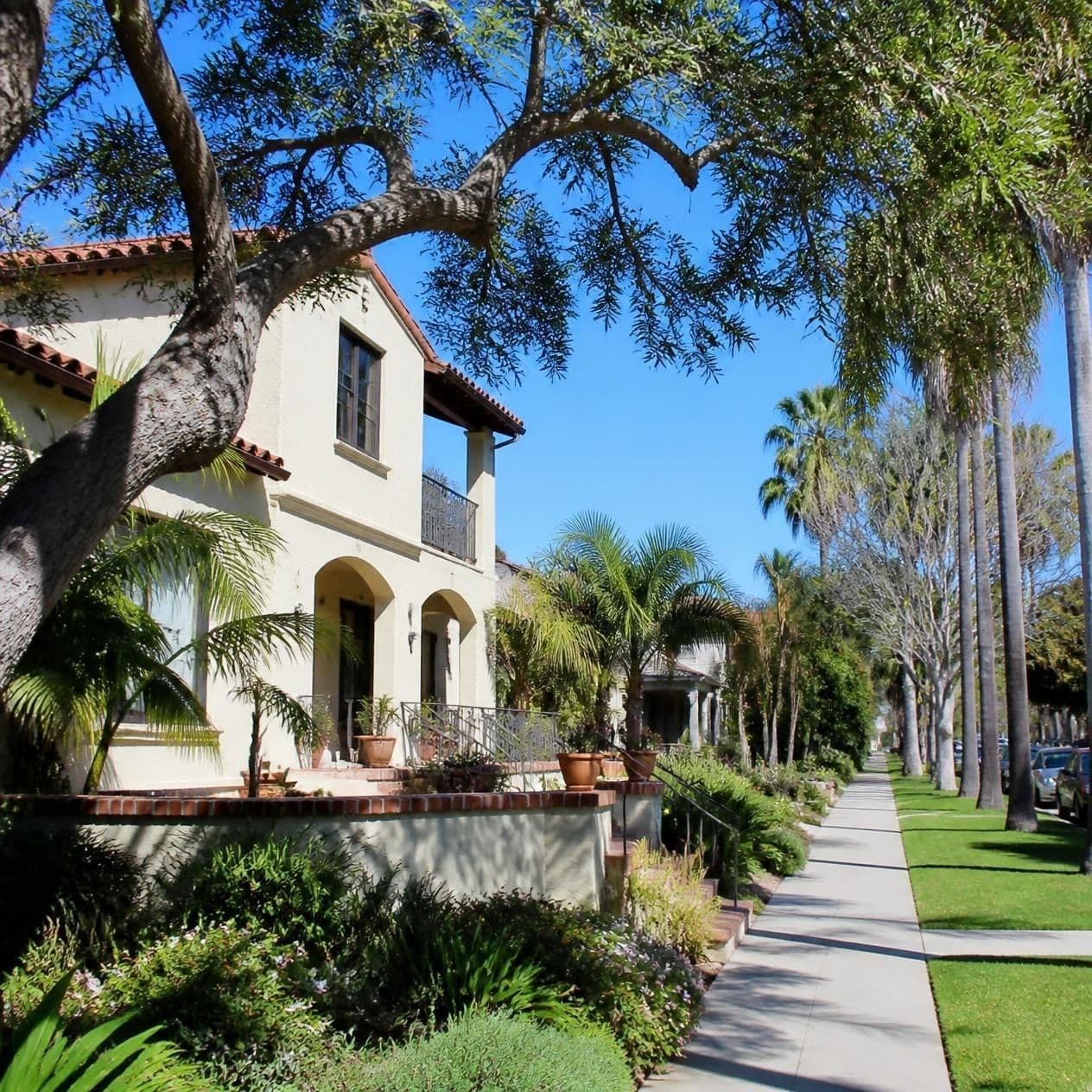 Mediterranean-style home with lush landscaping and palm trees along a sunny residential sidewalk.